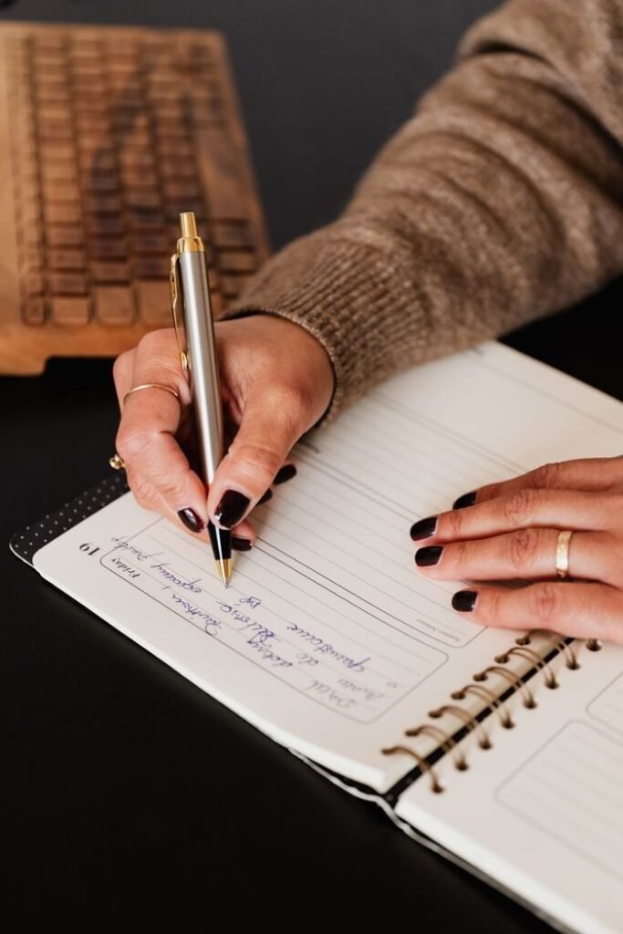 From above crop unrecognizable female with manicured hands taking notes in diary with stylish silver pen near computer keyboard